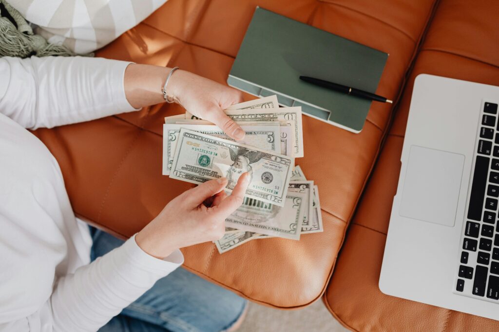 pexels-photo-5902918-5902918-1 Person holding US dollar bills on brown sofa beside laptop and notebook, representing financial management at home.